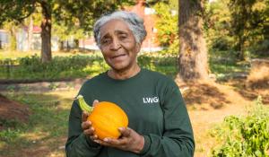 Cheryl Alston stands in a green long sleeve shirt holding a small pumpkin. She is looking calmly and pleasantly at the camera.