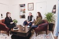 Athens Impact Socially Responsible Investments' founder Michelle Williams sits in a brown leather armchair - on her right are two clients, a white man in a black vest and a white woman with long brown hair in a green sweater, sitting in brown armchairs. Michelle has short hair, wears a black pantsuit, and has a folder in her hands.