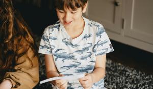 a boy of about ten years old reads a clue off a strip of paper. He is smiling and wearing a cotton t-shirt with alligators on it. His sister sits next to him, reading off the paper.