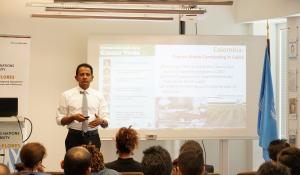 Hiroshan Hettiarachchi is giving a seminar, speaking to a class of diverse people that are all facing him. Behind Hettiarachchi is a powerpoint and to the far right is the UN flag.