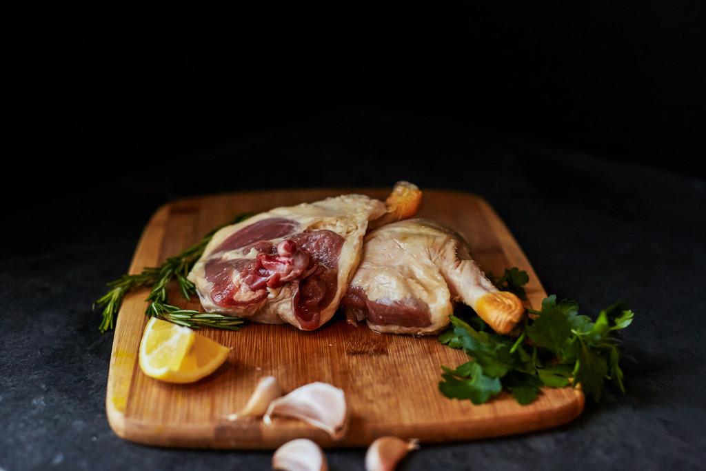 Uncooked duckling leg quarters on a wooden cutting board. There's a slice of lemon, garlic cloves, and herbs like rosemary and parsley. Sustainable Kitchen Products.