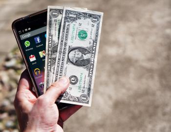 Image: hand holding smartphone and cash. Topic: What Does it Mean to Vote With Your Dollar?