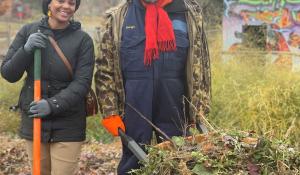 Michael Chaney is wearing a big brown coat and a red scarf, holding a wheelbarrow filled with foliage for compost. He stands next to Jasmira Colon who is smiling at the camera and holding a shovel with an orange handle.