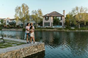 Two women stand with their arms around a young girl, their daughter, at the edge of a small lake with homes and trees in the background. Clean energy for all.