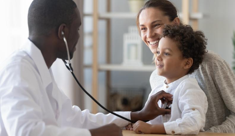A young black doctor holds a stethoscope up to a mixed child's heart. The mom has the child on her lap. Both are smiling at the doctor.