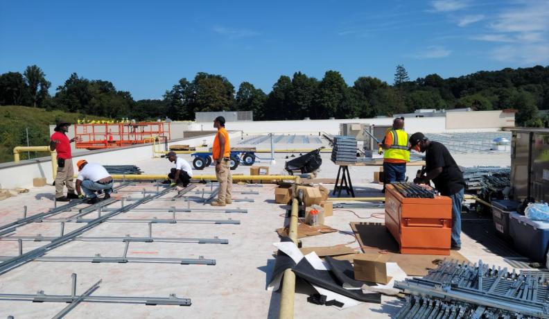 six solar workers standing on a rooftop installing solar panels. Each one is doing something different, whether drilling together metal pieces, supervising, or assembling the pieces together.