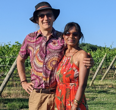 Swati & Mark standing together against a green landscape. Swati is wearing a bright orange dress and they are both smiling.