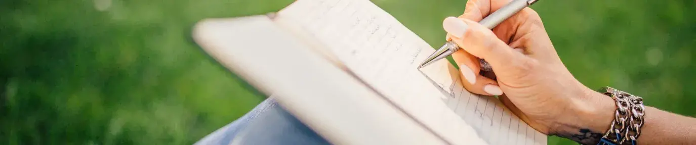 A woman's hand writes in a notebook resting on her knees. She's sitting in a grass field. Sustainable creative accessories.